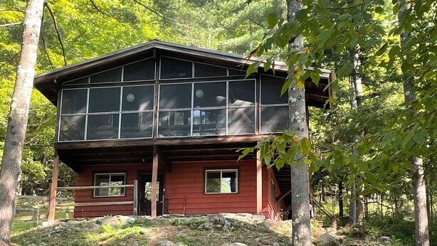 A cabin with a large windowed front section, surrounded by a grassy area with red Adirondack chairs. - Forest Hideaway (Keene)