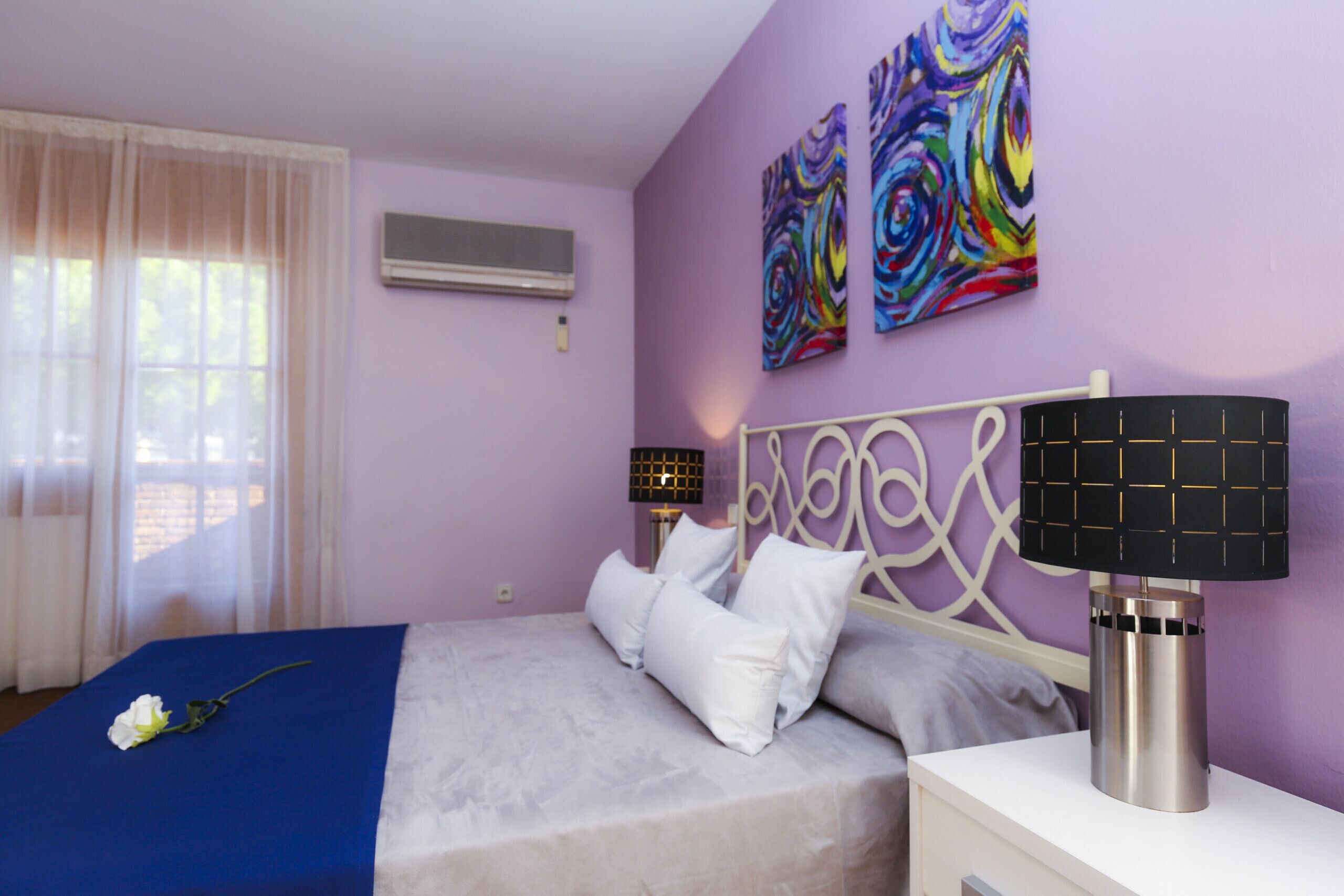 A bedroom with a bed, white pillows, a blue bedspread, a bedside lamp, and two abstract paintings on the wall.