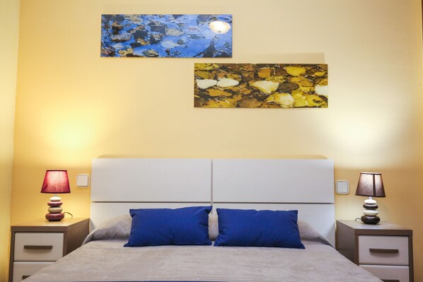Un dormitorio con una cama, dos almohadas azules, dos mesitas de noche con lámparas y dos cuadros en la pared.