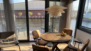 A dining area with a round table, six chairs, a woven light fixture, and a view of a water body and buildings through large windows.