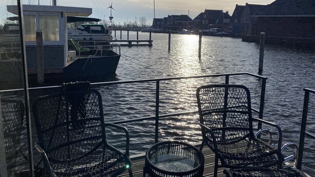 A boat deck with metal chairs and a small table, a dock with a boat, and buildings in the background.