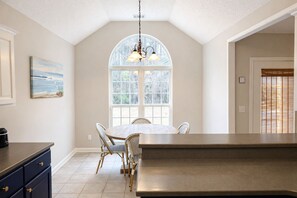 Een keuken met een groot eiland, een eettafel met stoelen, een hanglamp en een raam met uitzicht op bomen.