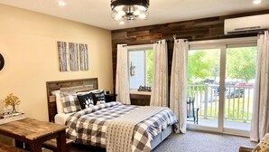 Un dormitorio con cama, una mesa de madera, un banco, una ventana con cortinas y un balcón con vistas a coches y vegetación.