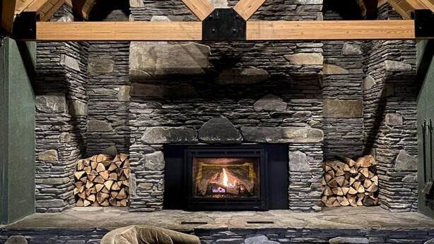 A stone fireplace with a fire, flanked by stacked firewood, in a room with a sloped ceiling and wooden beams.