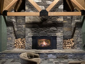 A stone fireplace with a fire, flanked by wooden logs, in a room with a stone wall and a wooden ceiling. - River & Loon Mountain View Studio - Indoor Pool & Hot Tub, Sleeps 3 (Lincoln)