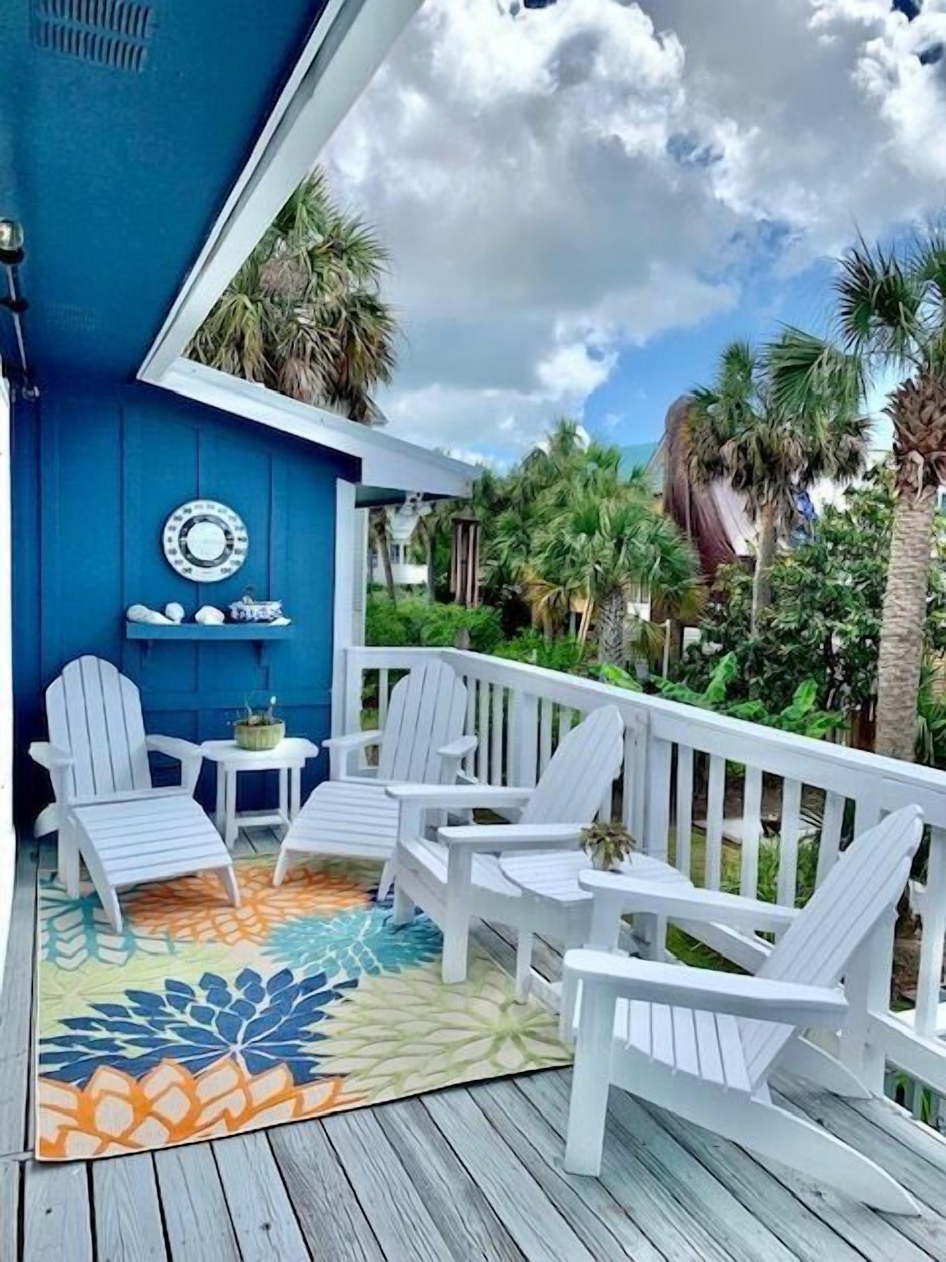 A blue porch with white Adirondack chairs and a small table, surrounded by lush greenery and palm trees.