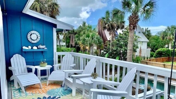 En terrass med vita Adirondack-stolar och en färgglad matta, med utsikt över en pool och frodig grönska.