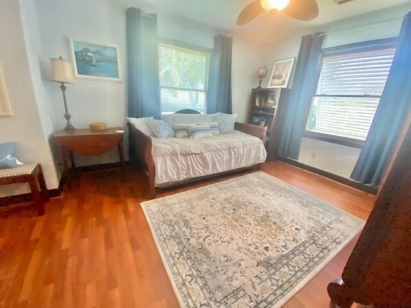 A bedroom with a bed, a nightstand, a chair, a rug, a window with curtains, and a ceiling fan.