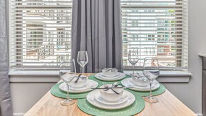 A dining table set for four with white plates, green placemats, and wine glasses, positioned by a window with blinds.