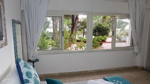 Een slaapkamer met een bed, kussens, een raam met uitzicht op groen en een klein tafeltje met een lamp.