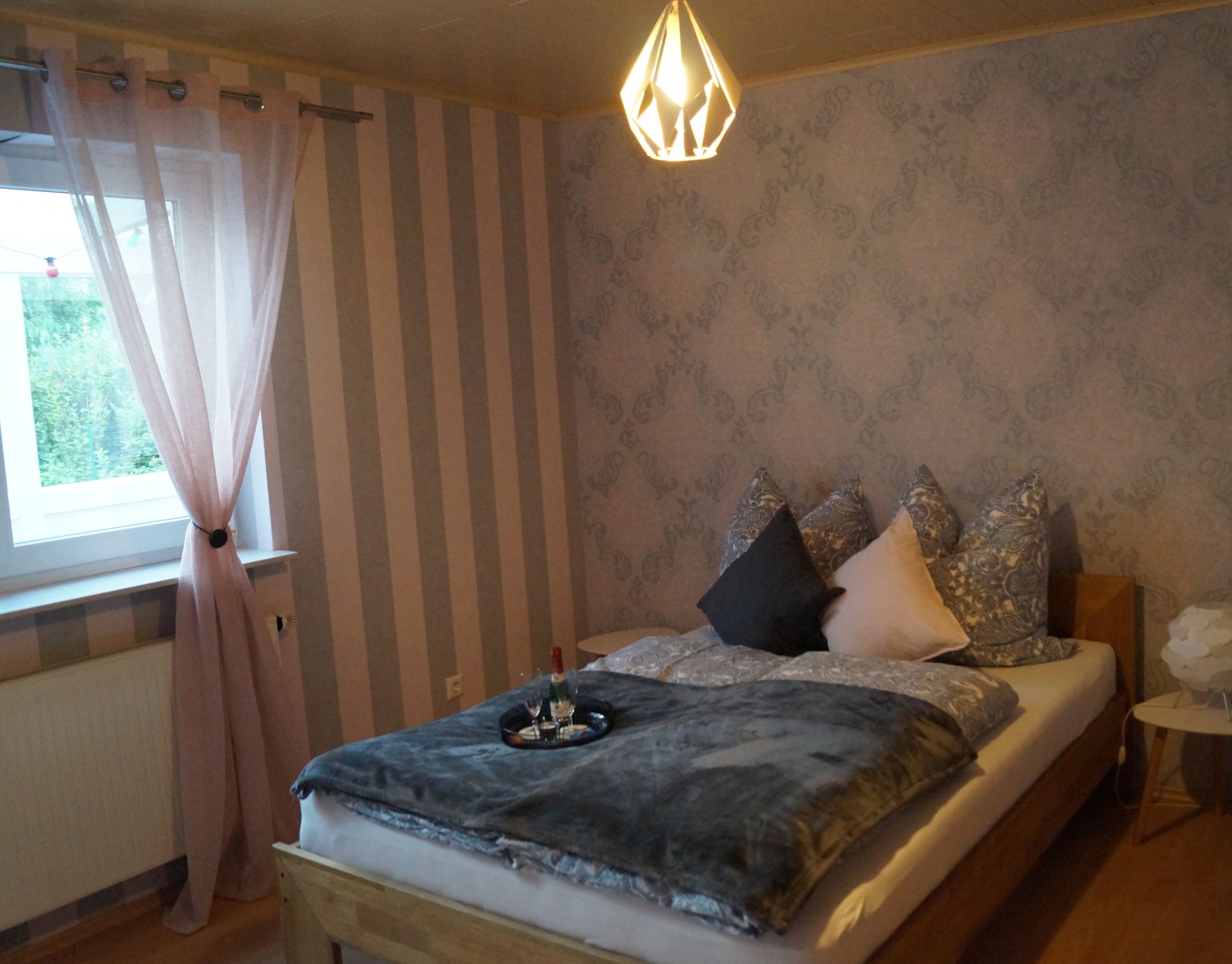 Une chambre à coucher avec un lit, des rideaux, un luminaire suspendu, une petite table et un mur orné d’un motif.
