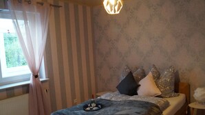 A bedroom with a bed, curtains, a hanging light fixture, a small table, and a wall with a pattern. - Quietly located vacation apartment in Reichshof Eckenhagen (Reichshof)