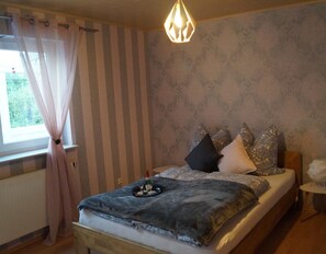 Une chambre à coucher avec un lit, des rideaux, un luminaire suspendu, une petite table et un mur orné d’un motif.