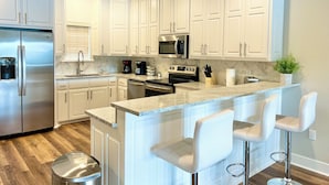 A modern kitchen with white cabinetry, stainless steel appliances, a marble countertop, and a bar area with white stools. - Stylish New Lake Condo with180° Lakefront View- Main Channel, Central Location! (Hot Springs)