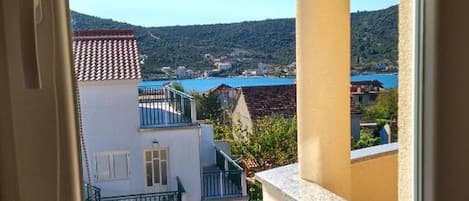 A window with a view of a coastal town, a balcony, and a vase with dried flowers on the windowsill.
