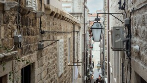 Une ruelle étroite pavée, bordée de bâtiments historiques en pierre, dotés de fenêtres à volets et d’espaces aménagés pour s’asseoir à l’extérieur.