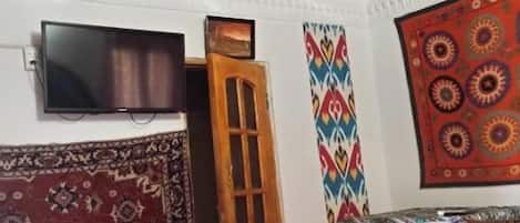 A room with a wooden ceiling, a television mounted on the wall, a patterned rug, a colorful wall hanging, and a small table.