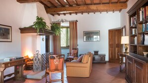 A cozy living room with a wooden ceiling, a fireplace, a sofa, a bookshelf, and a dining table. - Villa Eama - Tuscan Villa with Private Pool (Pian di Scò)