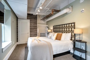 A hotel room with a bed, a nightstand, a lamp, a window, and a door.