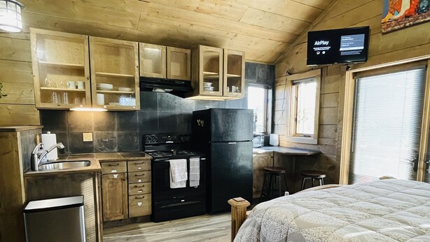 A compact kitchen with wooden cabinets, a black refrigerator, a stove, and a sink. There is a television mounted on the wall and a window with blinds. - Historic Westcliffe Outpost Loft (Westcliffe)