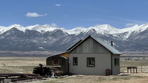 A small, single-story building with a sloped roof, situated in a rural area with a backdrop of snow-capped mountains. - Historic Westcliffe Outpost Loft (Westcliffe)