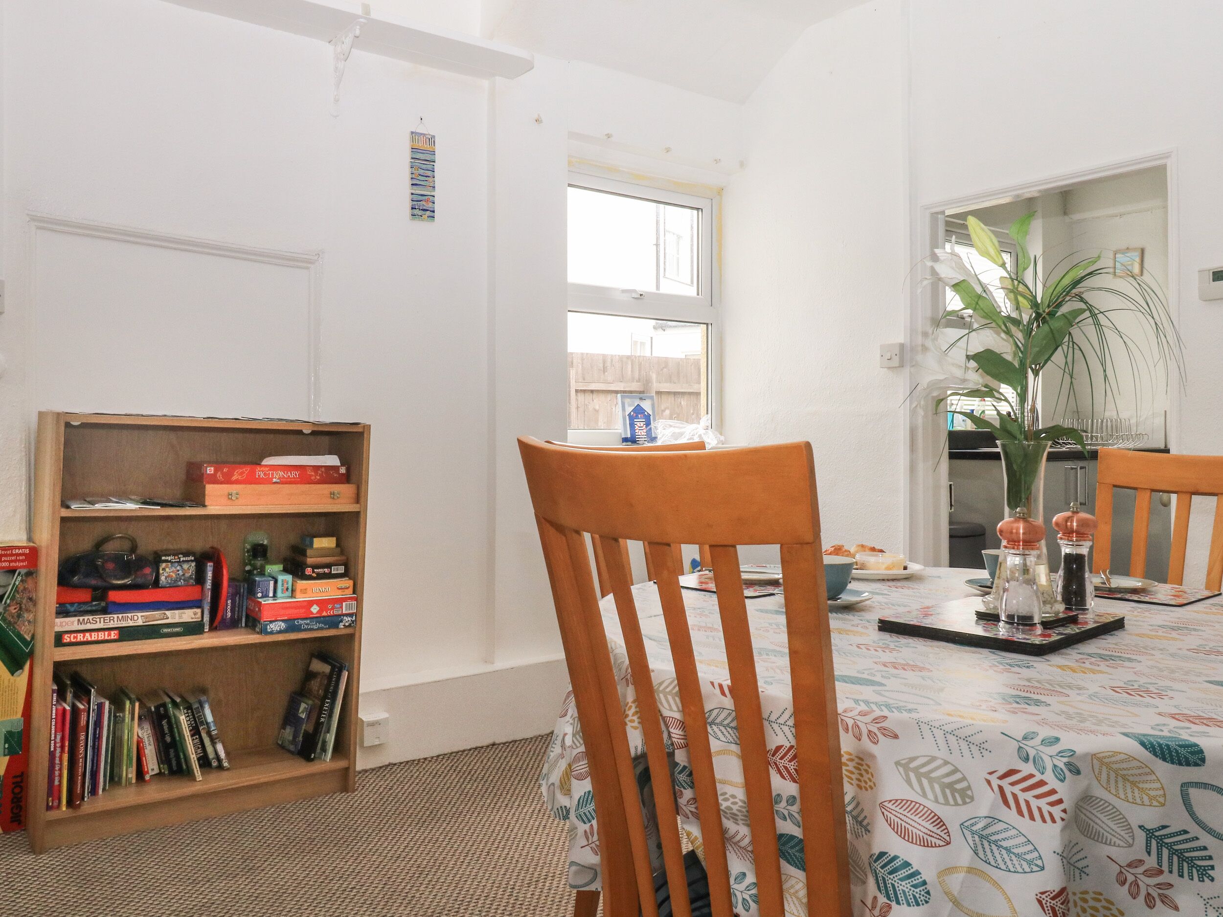 A dining area with a wooden bookshelf, a table with a patterned tablecloth, and a window with a view of a wooden fence.