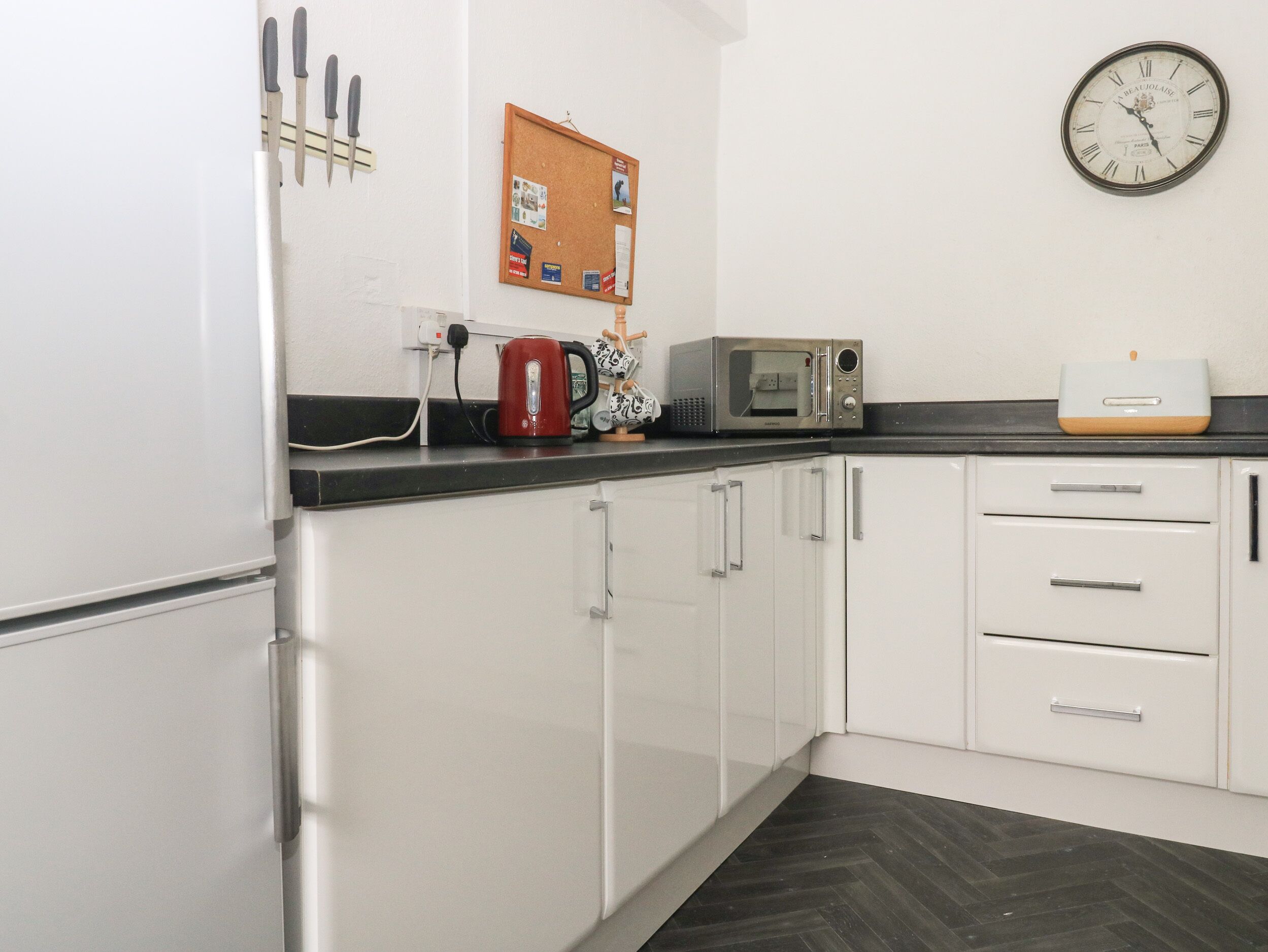 A kitchen with white cabinets, a microwave, a kettle, and a clock on the wall.