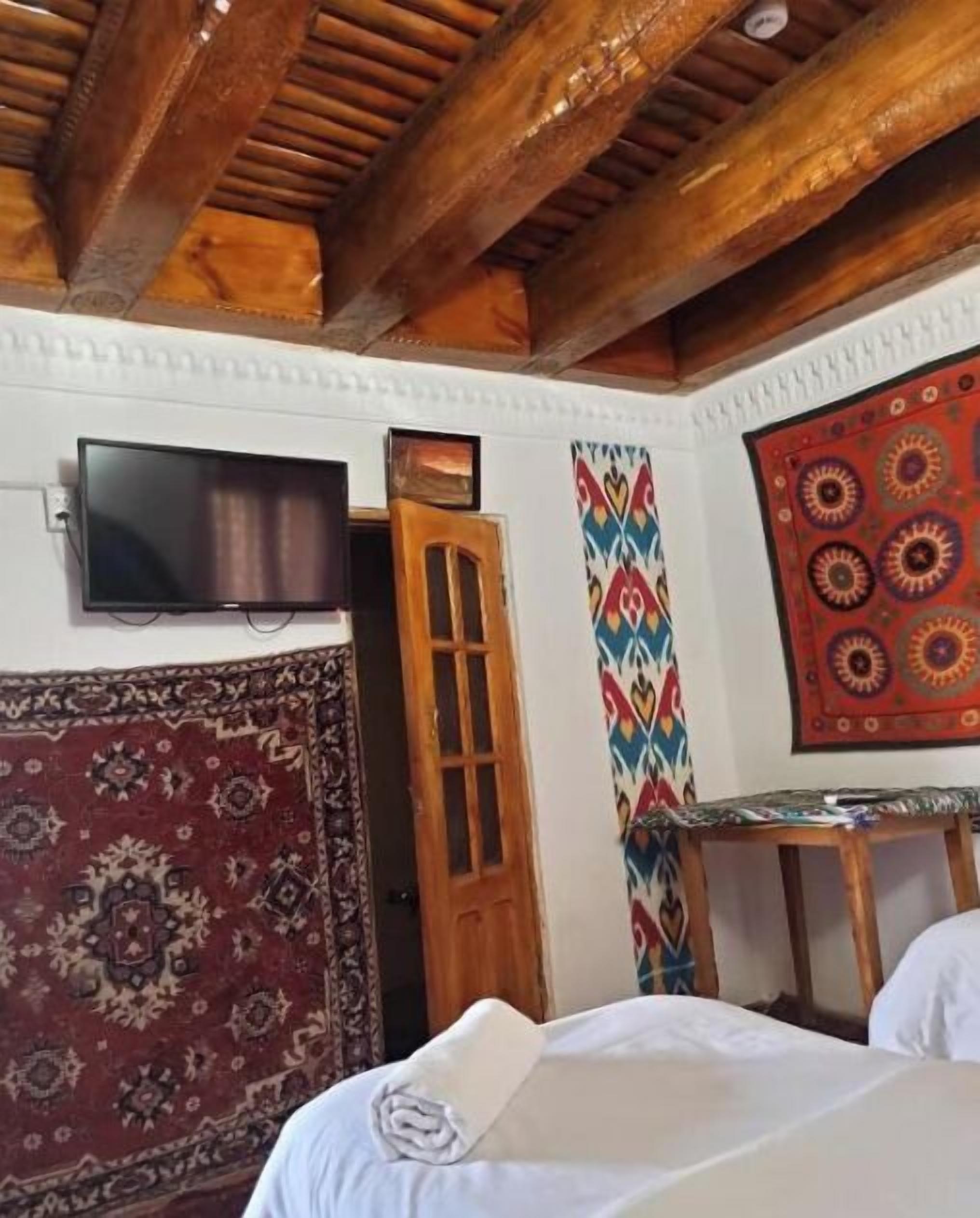 A room with a wooden ceiling, a television mounted on the wall, a patterned rug, a colorful wall hanging, and a small table.