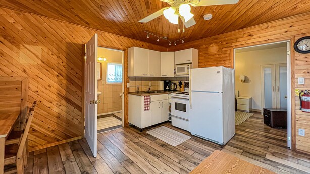 A compact kitchen with wooden walls and a ceiling fan, a small dining area, and a visible clock on the wall. - Ahmic Lake Resort's 1-Bedroom Cottage w/ hot tub (LOON Cottage) (Magnetawan)