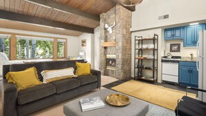 A modern living room with a stone fireplace, a grey sofa with yellow cushions, and a kitchen with blue cabinets and a white stove. - Aspenwood by Snowmass Vacations (Snowmass Village)