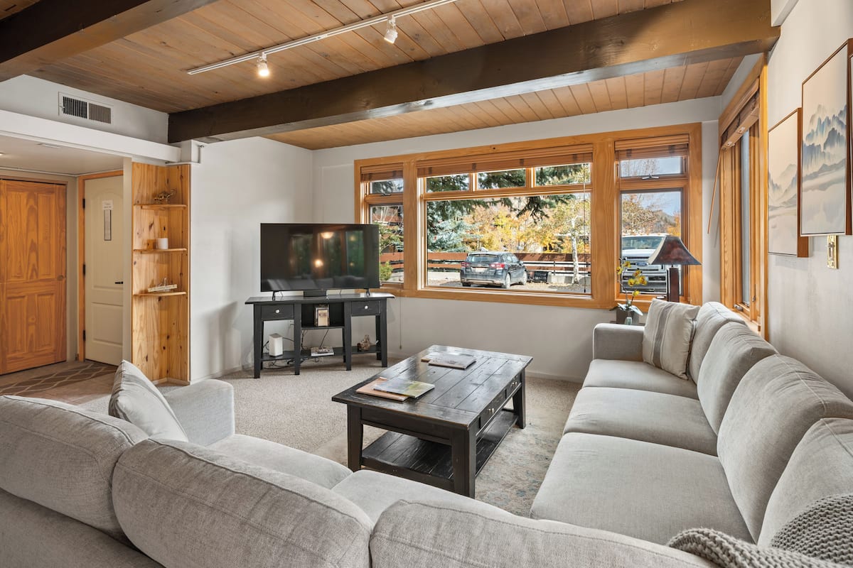 Una sala de estar moderna con un sofá grande, una mesa de centro y un televisor. Se observa un mueble de madera y una ventana con vistas al exterior.