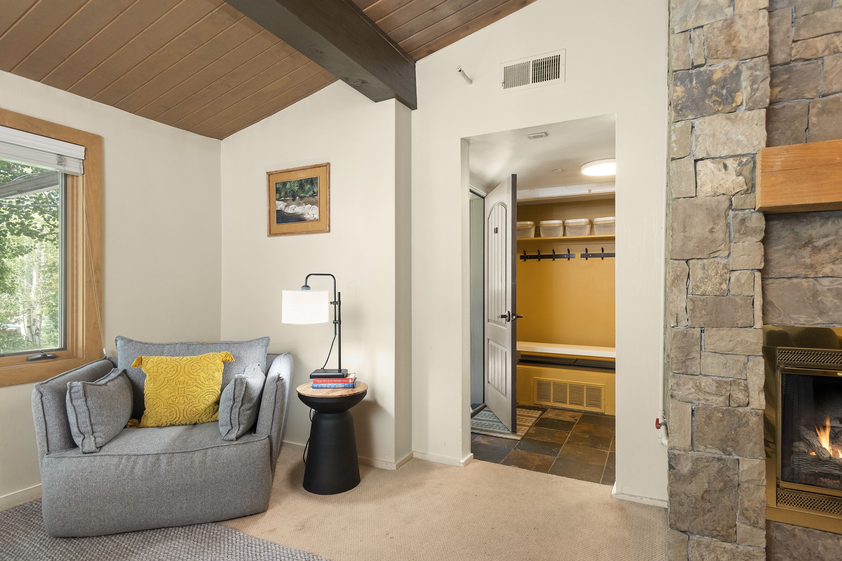 A cozy living room with a gray sofa, a small side table with books, a lamp, and a stone fireplace.