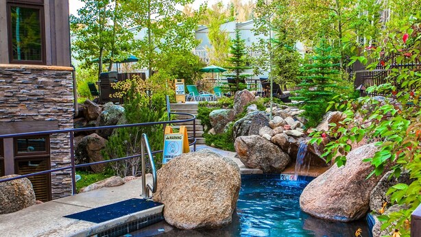 A pool area with large rocks, a warning sign, and a view of a building in the background. - Aspenwood by Snowmass Vacations (Snowmass Village)