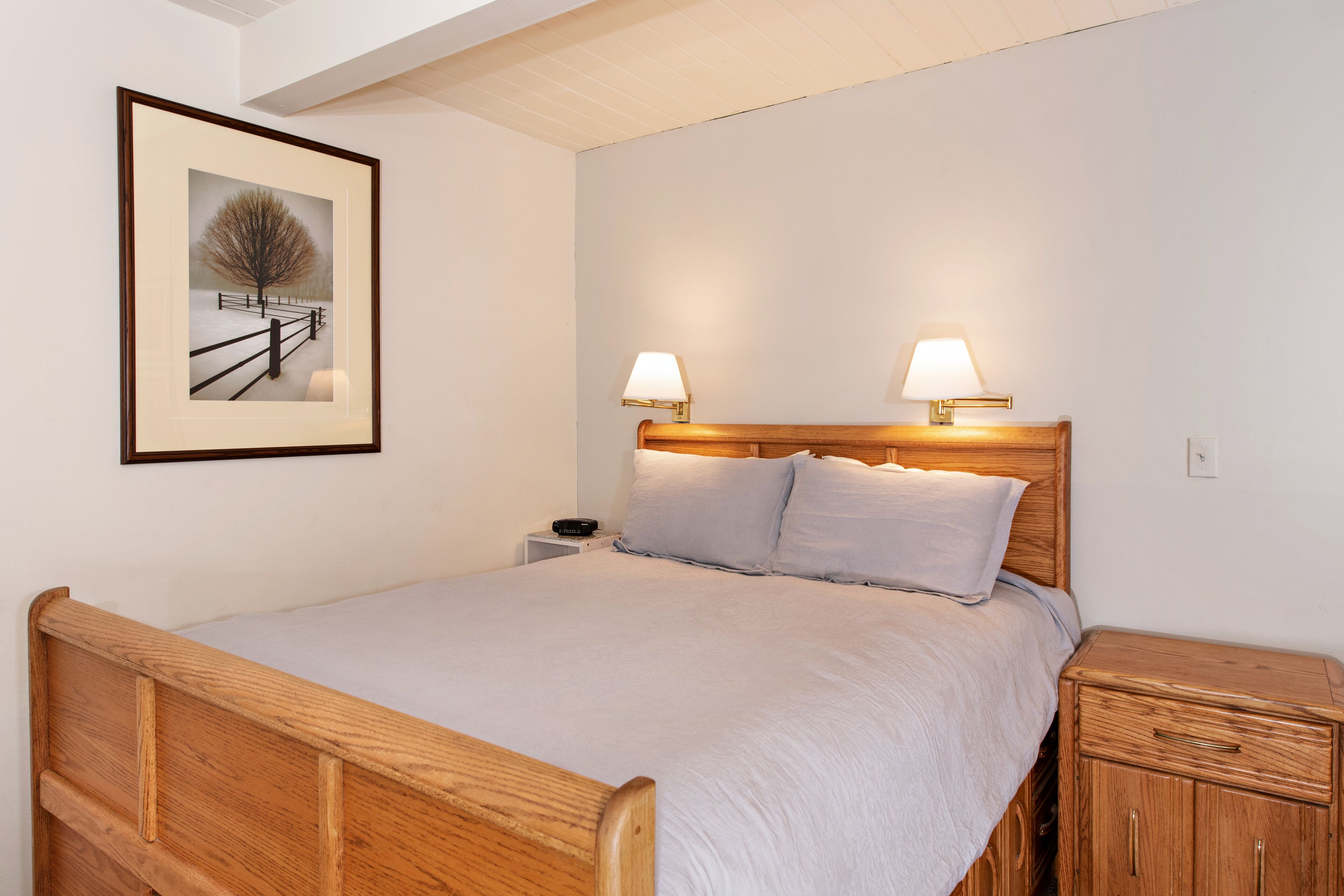 A neatly made bed with a wooden headboard, two bedside lamps, a nightstand, and a framed picture on the wall.