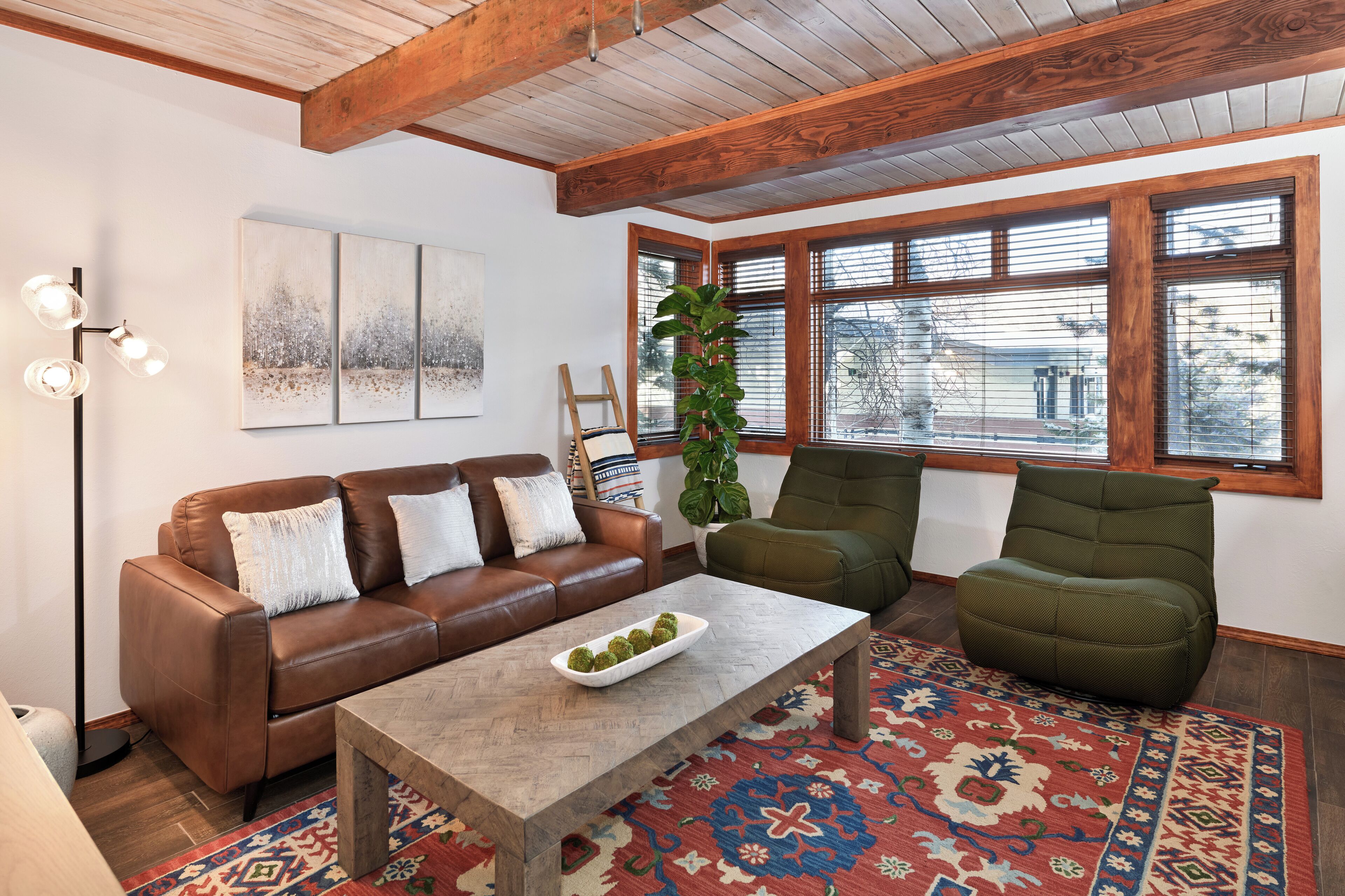 A living room with a brown leather sofa, a green armchair, a coffee table with decorative items, a patterned rug, and a large window with blinds.