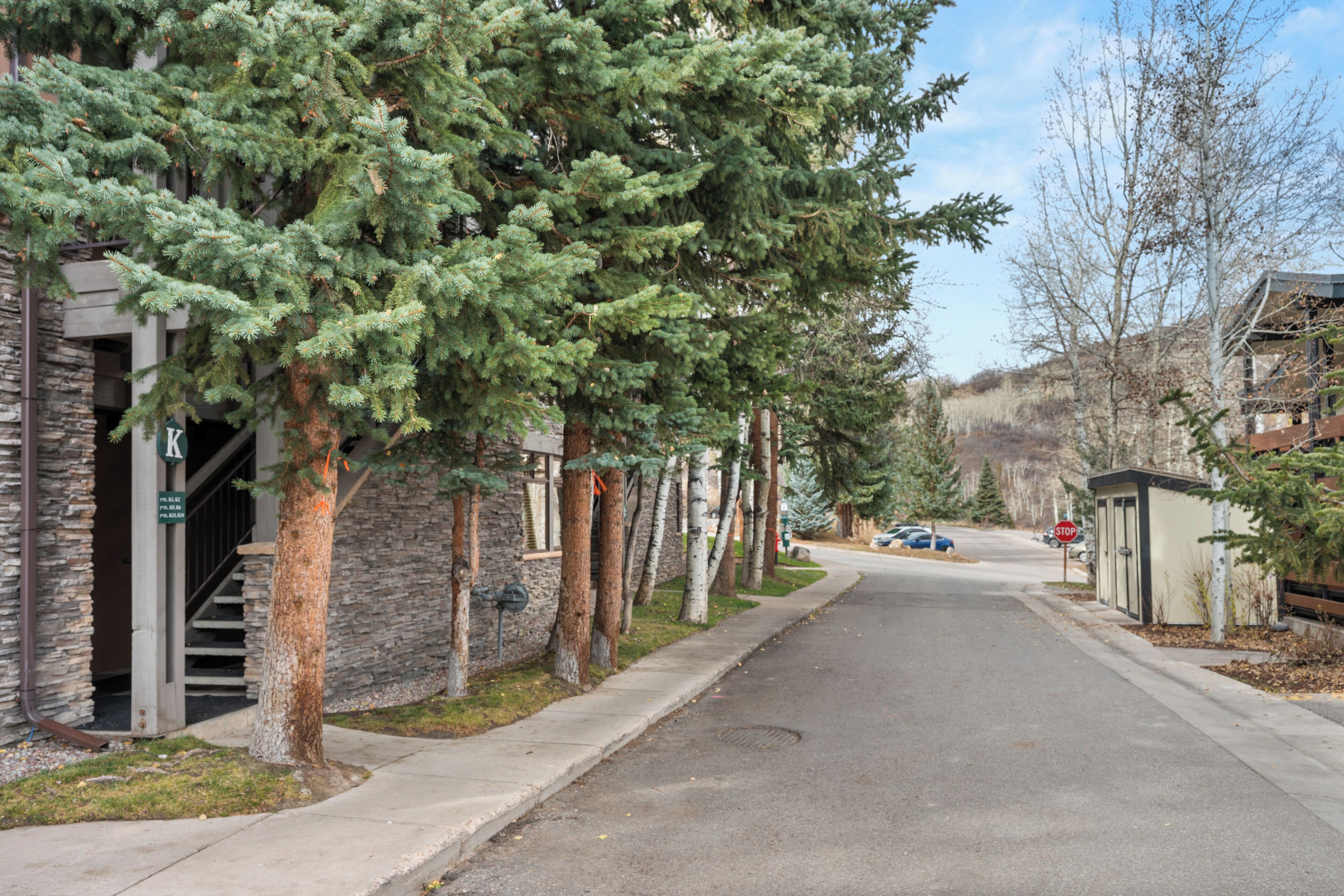 A street lined with trees and buildings, including a stone building with a visible staircase and a small structure with a roof.