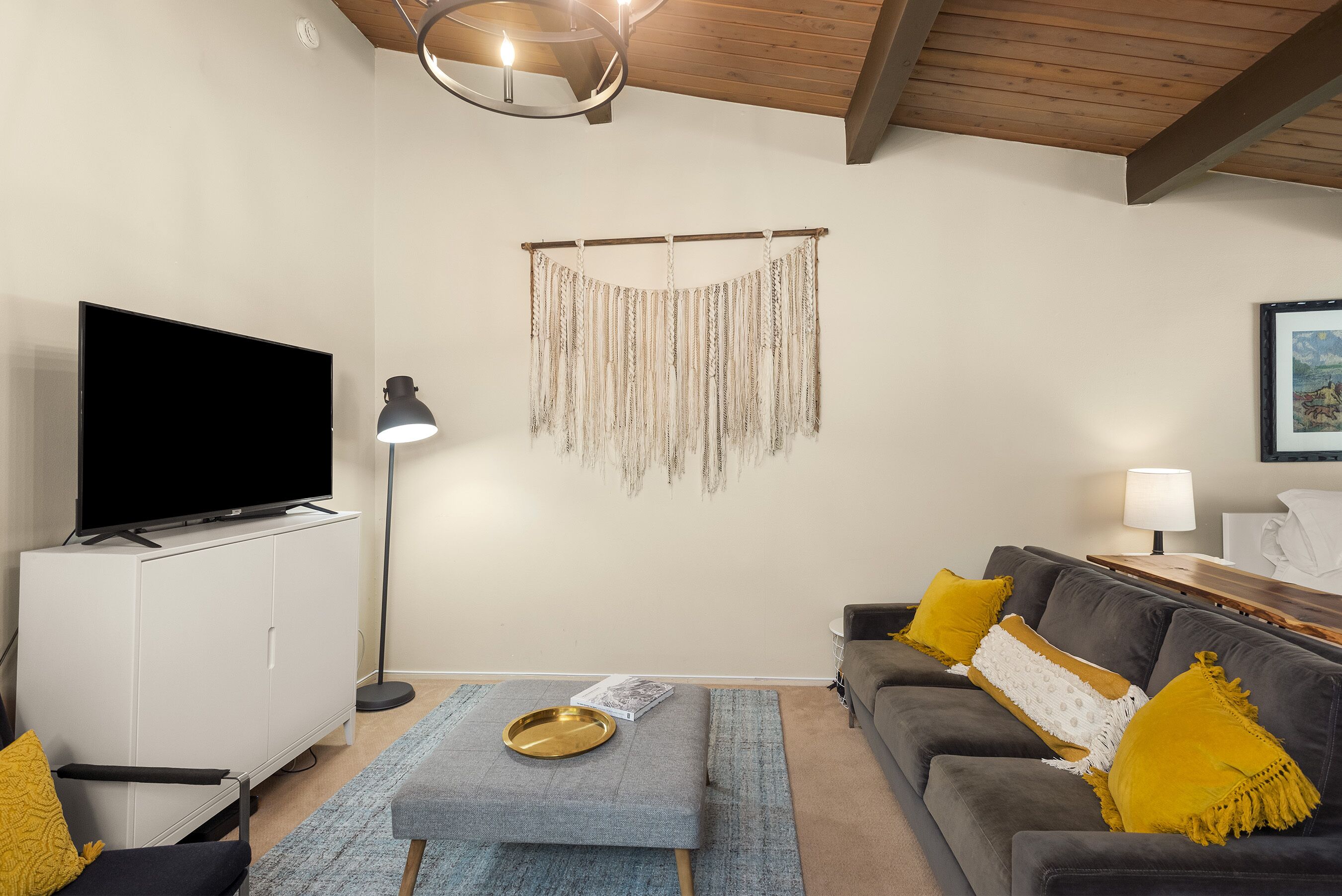 Una sala de estar con un televisor de pantalla plana, un mueble blanco, un sofá gris con cojines amarillos, una alfombra azul y un adorno en la pared.
