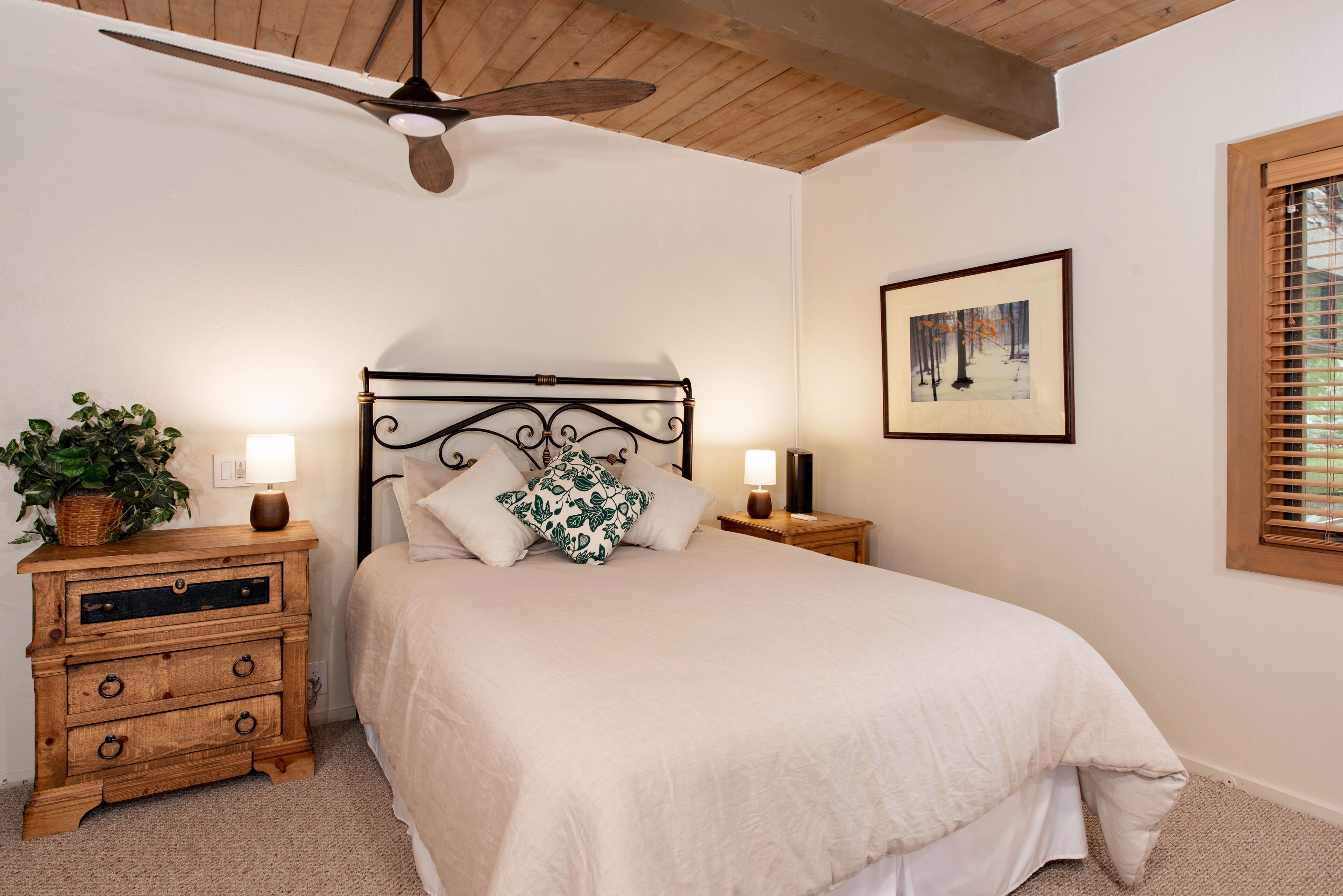 A bedroom with a bed, wooden nightstands, a ceiling fan, a framed picture on the wall, and a window with blinds.