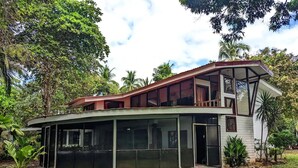 Ein modernes, zweistöckiges Haus mit großen Fenstern, einer überdachten Terrasse und einem Garten mit tropischen Pflanzen.