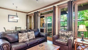 A living room with a leather sofa, armchair, and coffee table, large windows with curtains, and a dining area with chairs and a table.