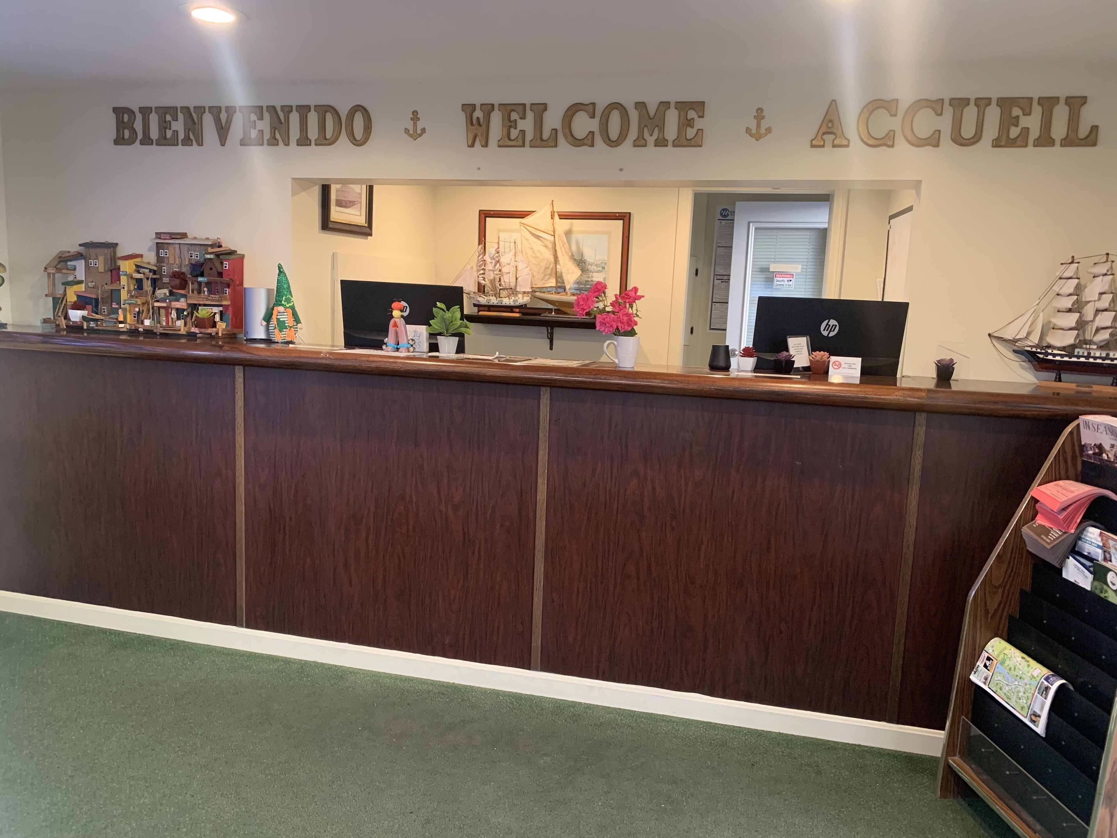 Un comptoir d'accueil avec un revêtement en bois, une enseigne indiquant « BIENVENIDO WELCOME ACCUEIL » au-dessus, et divers objets décoratifs, notamment un modèle de navire, des fleurs et un petit sapin de Noël.