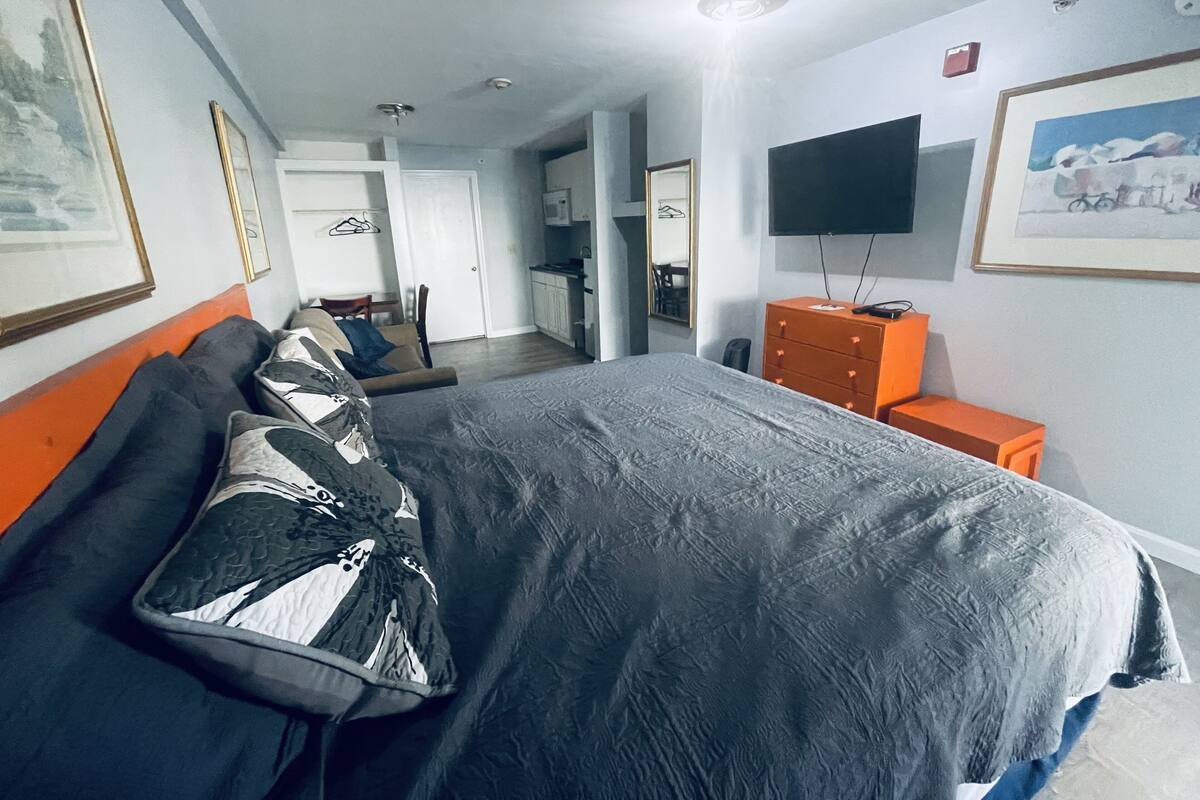 A hotel room with a large bed, a TV on the wall, and a kitchenette in the background.
