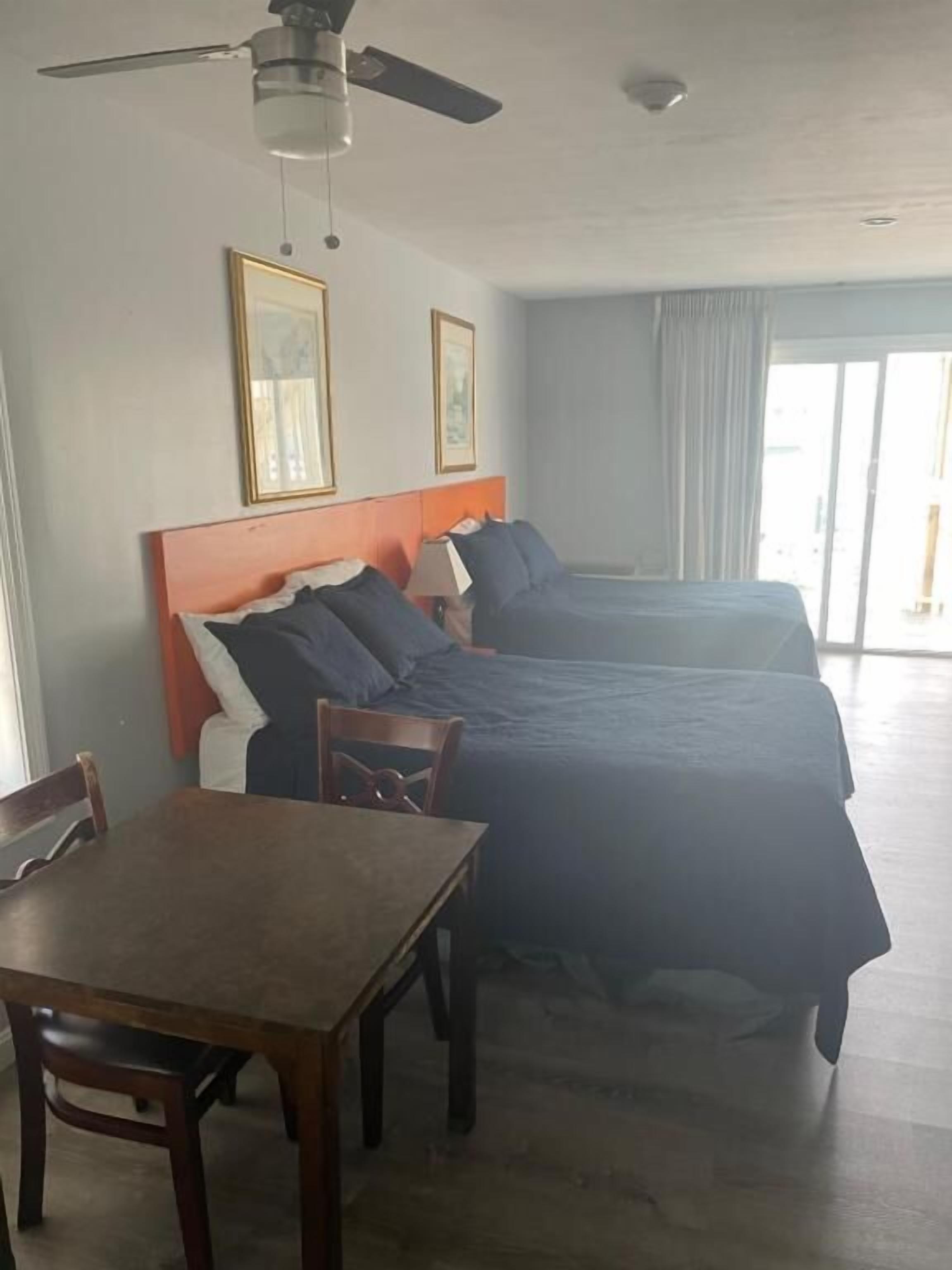 A hotel room with two beds, a dining table, chairs, ceiling fan, and large windows.