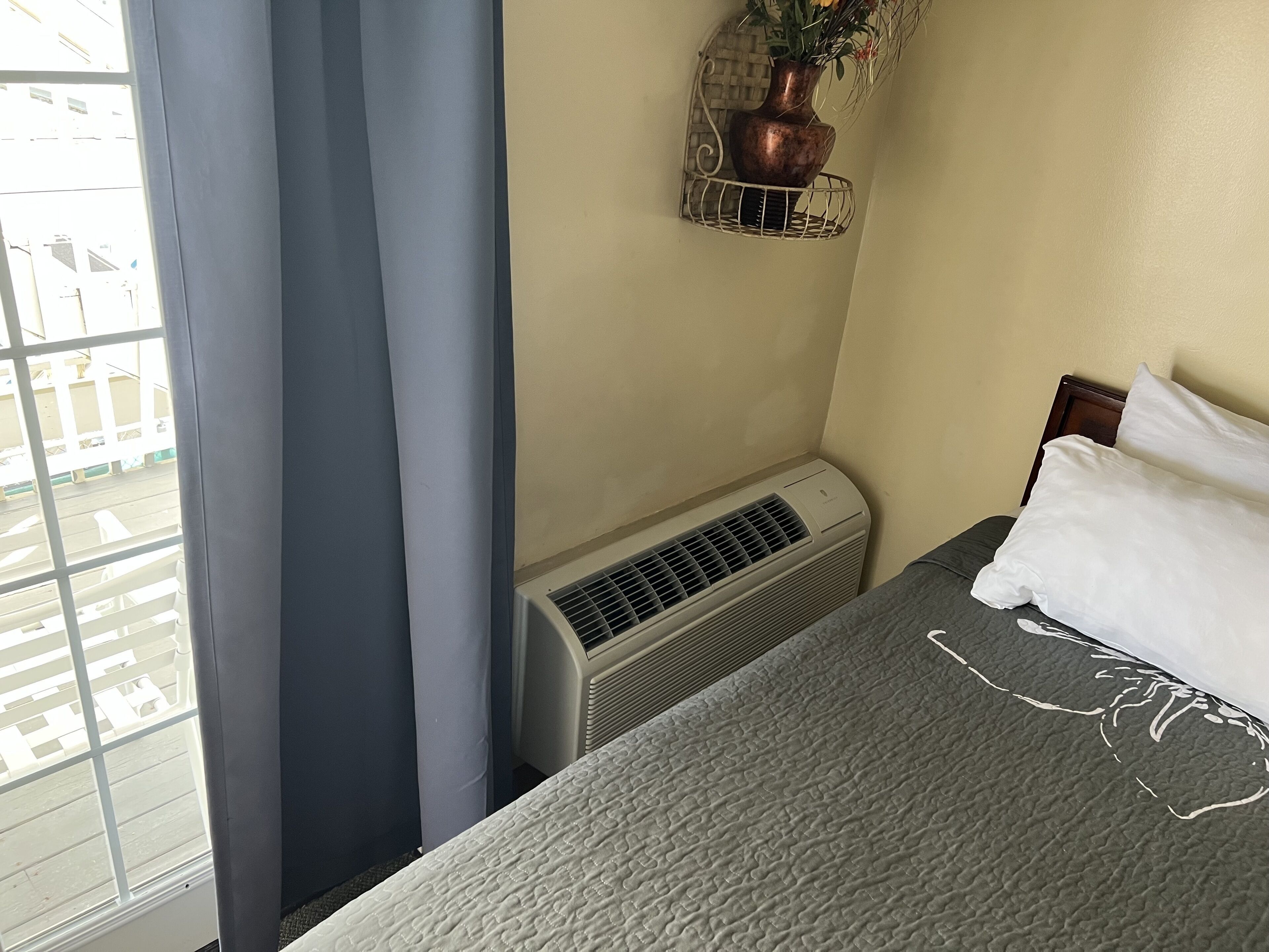 A hotel room with a bed, a window with curtains, an air conditioning unit, and a wall-mounted plant.