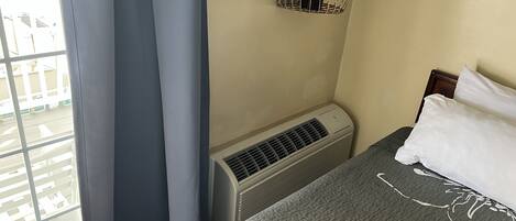 A hotel room with a bed, a window with curtains, an air conditioning unit, and a wall-mounted plant.