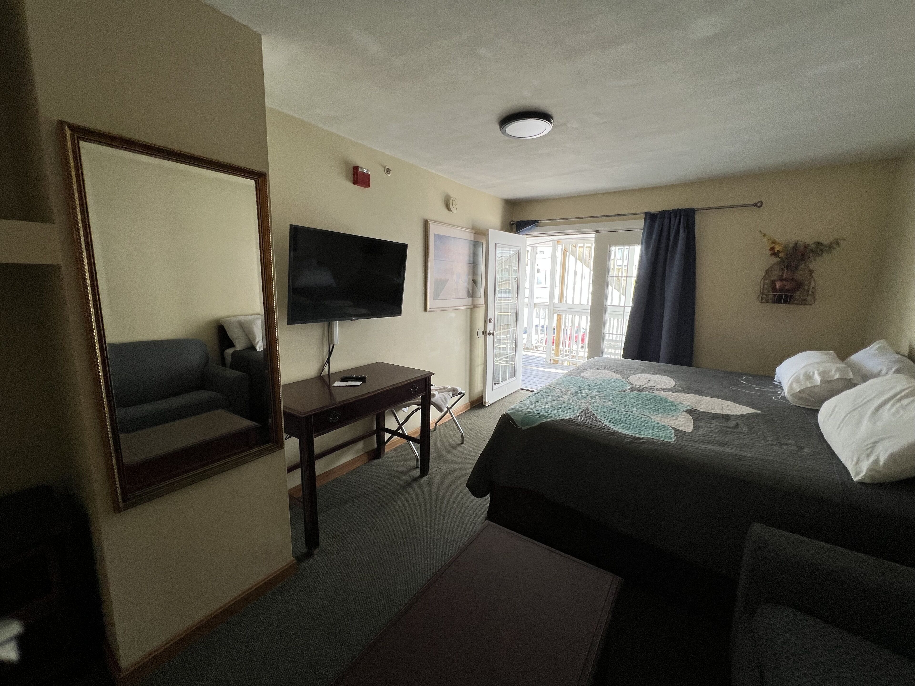 A hotel room with a bed, a desk with a TV, a chair, and a large mirror.