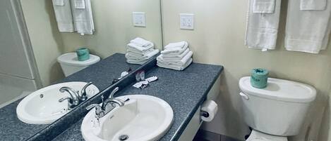 A bathroom with two sinks, a toilet, and a mirror. There are towels hanging on the wall and a toilet paper holder.