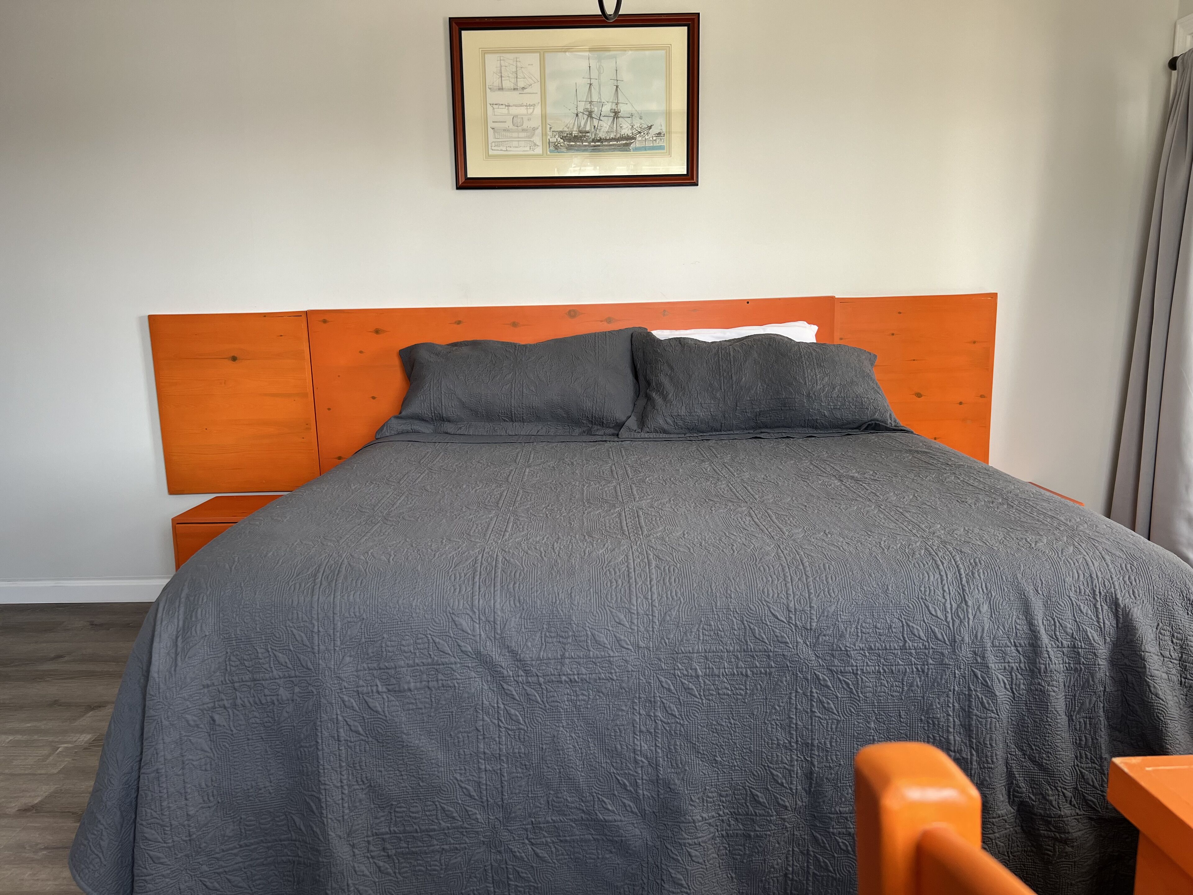 A bed with a grey quilt, wooden headboard, and a framed picture above it.