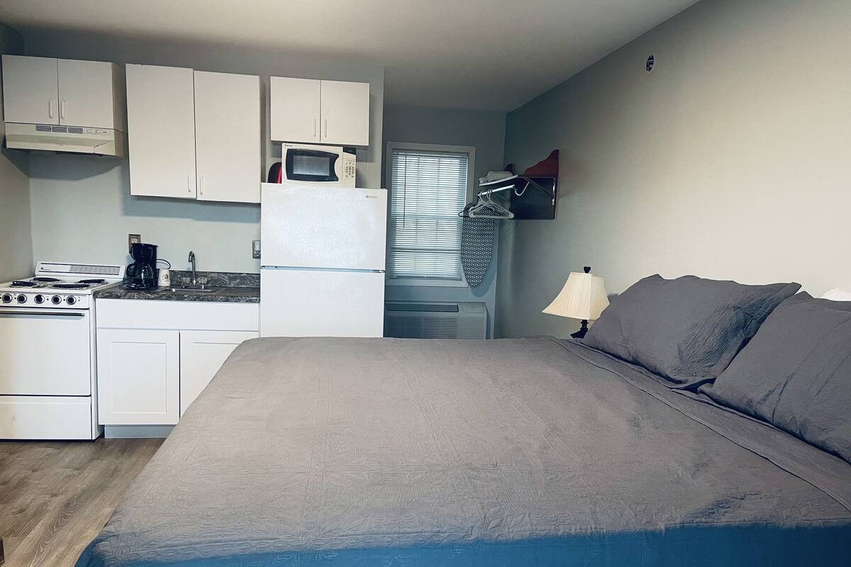 A compact room with a kitchenette, a bed, and a window with blinds.