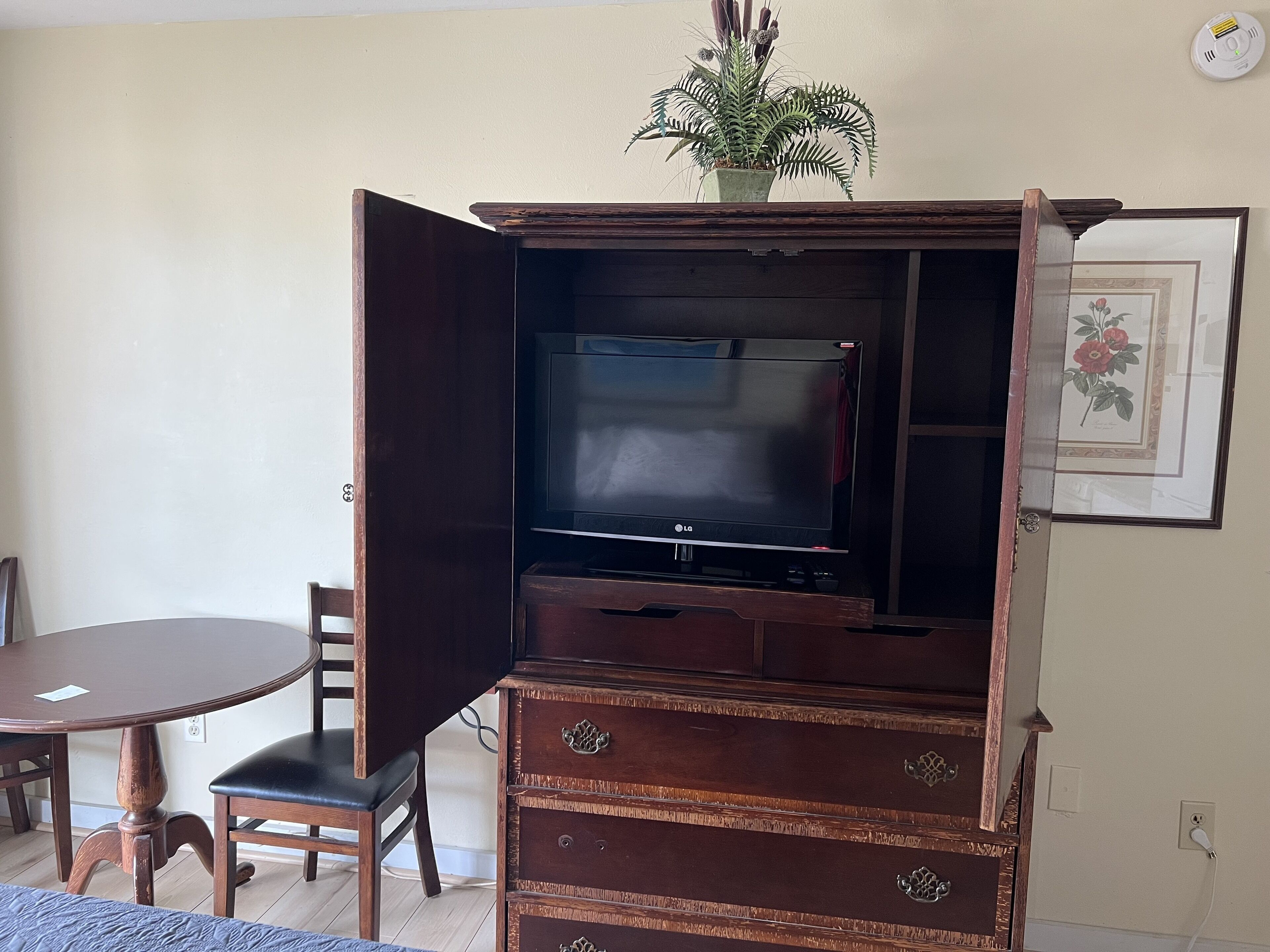 A living room with a TV on a wooden cabinet, a dining table with chairs, and a potted plant on top of the cabinet.